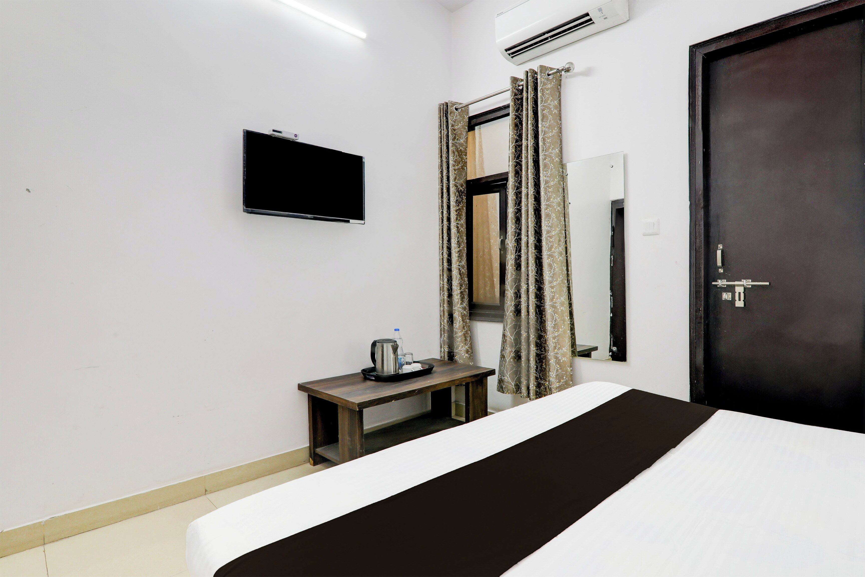 Une chambre d’hôtel avec un lit, une petite table avec une bouilloire et un plateau, un téléviseur fixé au mur et une porte avec un miroir.