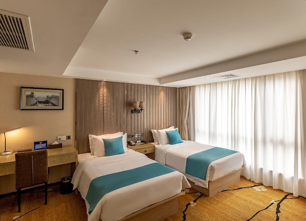Ein Hotelzimmer mit zwei Betten, einem Schreibtisch, einem Stuhl, einer Lampe, einem Bild an der Wand und einem Fenster mit Vorhängen.