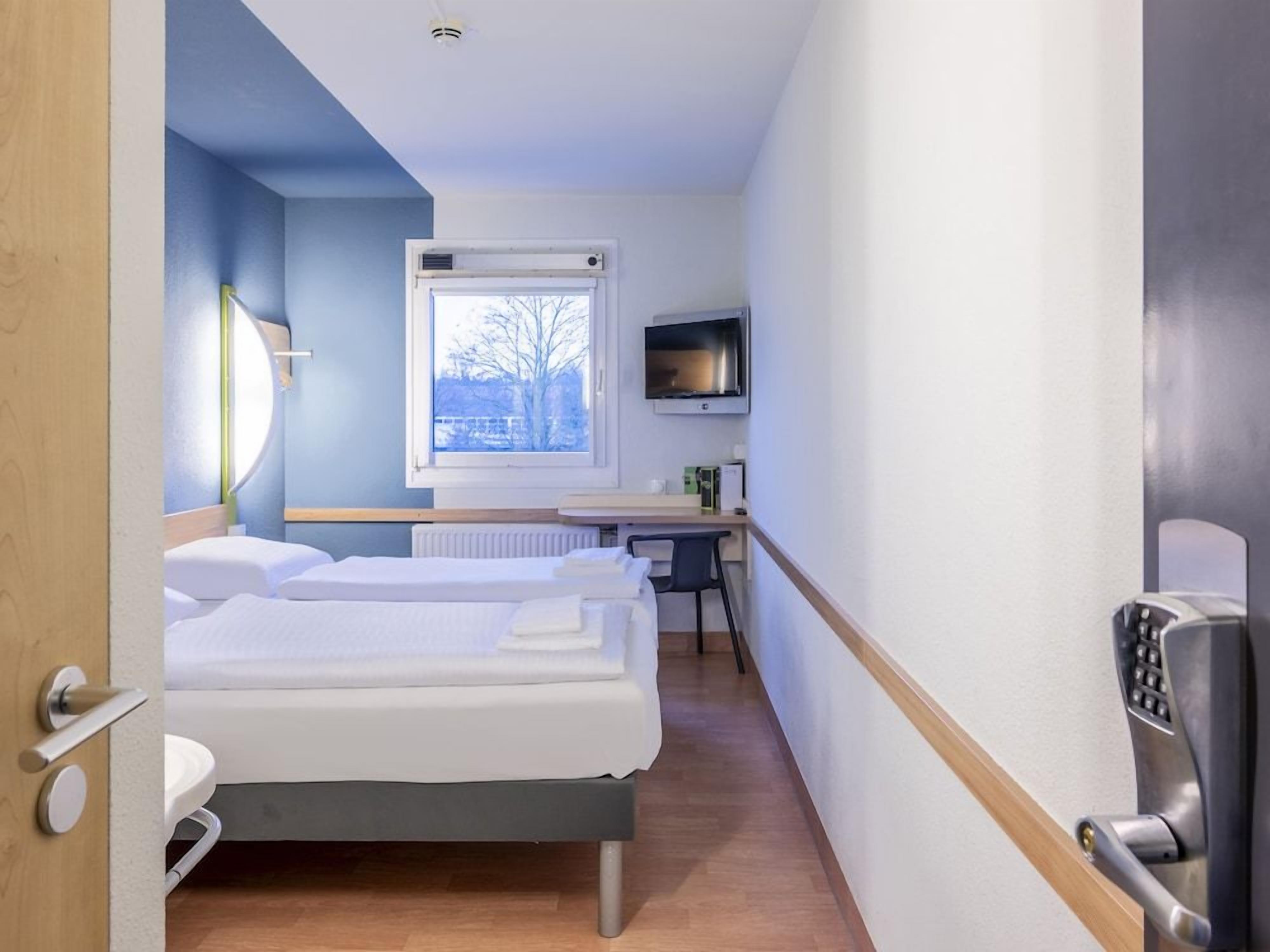 A hotel room with a bed, a desk with a chair, a television, and a window with a view of trees.