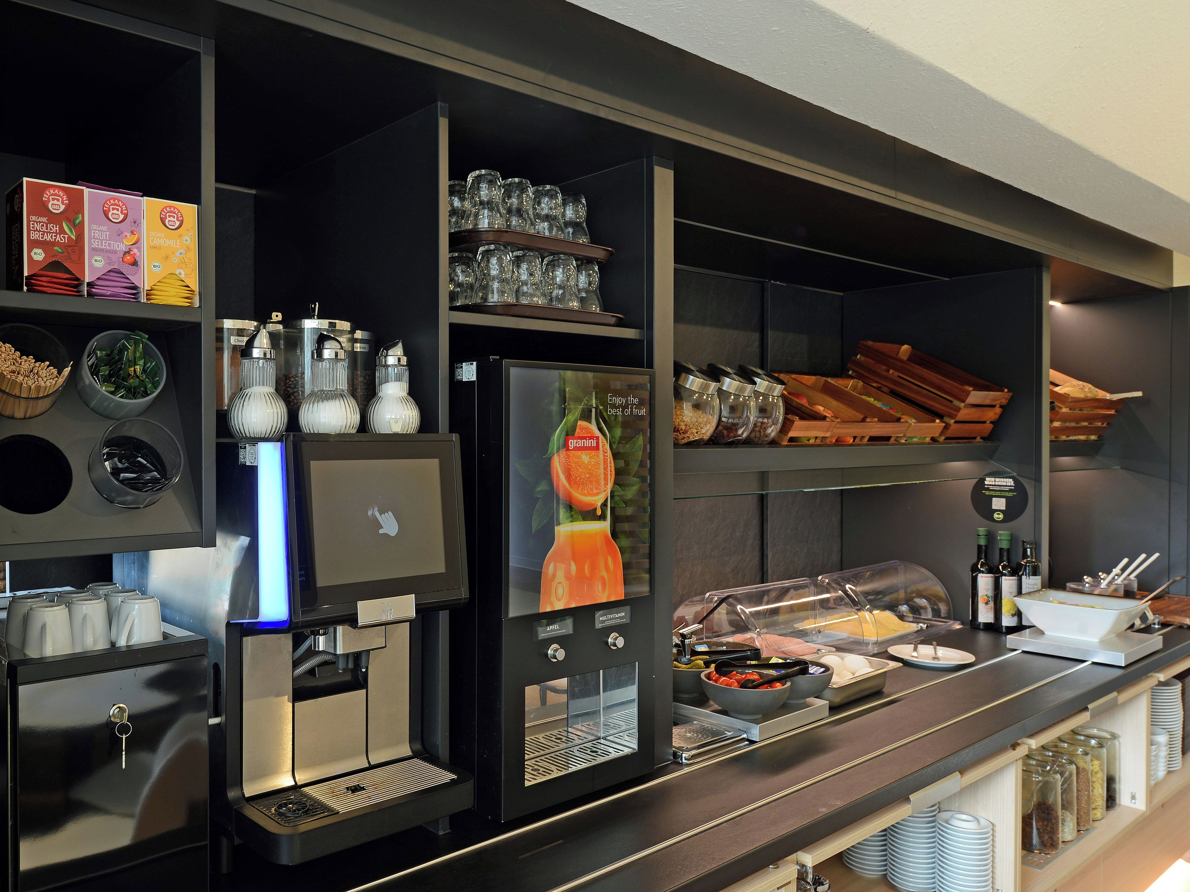 A self-service breakfast station with a coffee machine, various food containers, and a display of breakfast items.