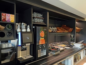A self-service breakfast station with a coffee machine, various food containers, and a display of breakfast items. - Ibis Budget Duesseldorf Willich (Willich)
