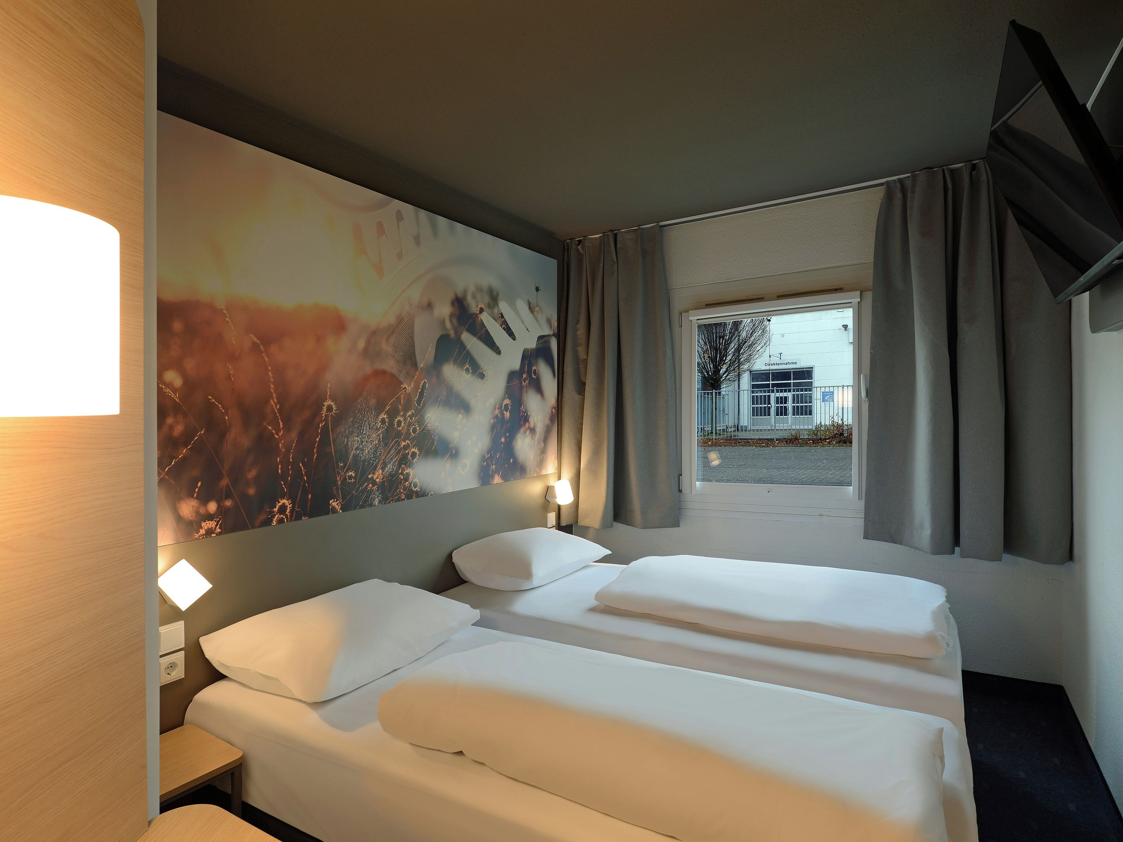 A hotel room with a bed, a nightstand, a wall-mounted lamp, and a window with curtains.