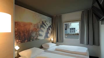 A hotel room with a bed, a nightstand, a wall-mounted lamp, and a window with curtains.