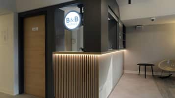 A reception desk with a B&B Hotels sign, a private room door, and a small table with a chair.