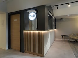 Una recepción con el cartel de B&B Hotels, una puerta de habitación privada y una pequeña mesa con una silla.