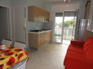 A compact kitchen with a gas stove, a dining table with a floral tablecloth, and a red sofa. - Central Apartment With Parking (San Michele al Tagliamento)