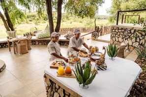 A dining area with a stone wall, wicker furniture, and a buffet setup. - Last Word Kitara Camp (Mpumalanga)