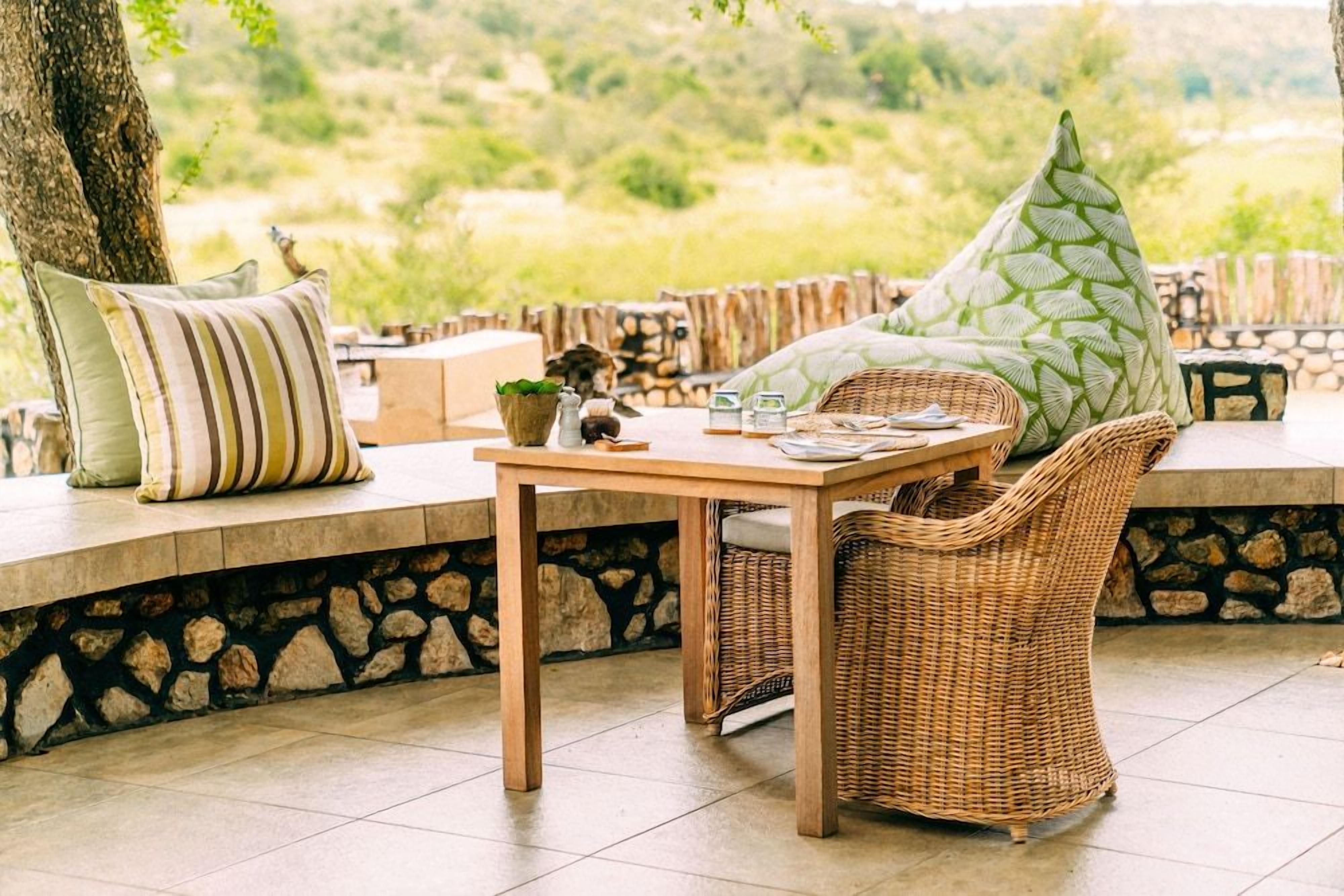 Une table en bois, des chaises et des coussins, un mur de pierre et une vue sur la verdure.