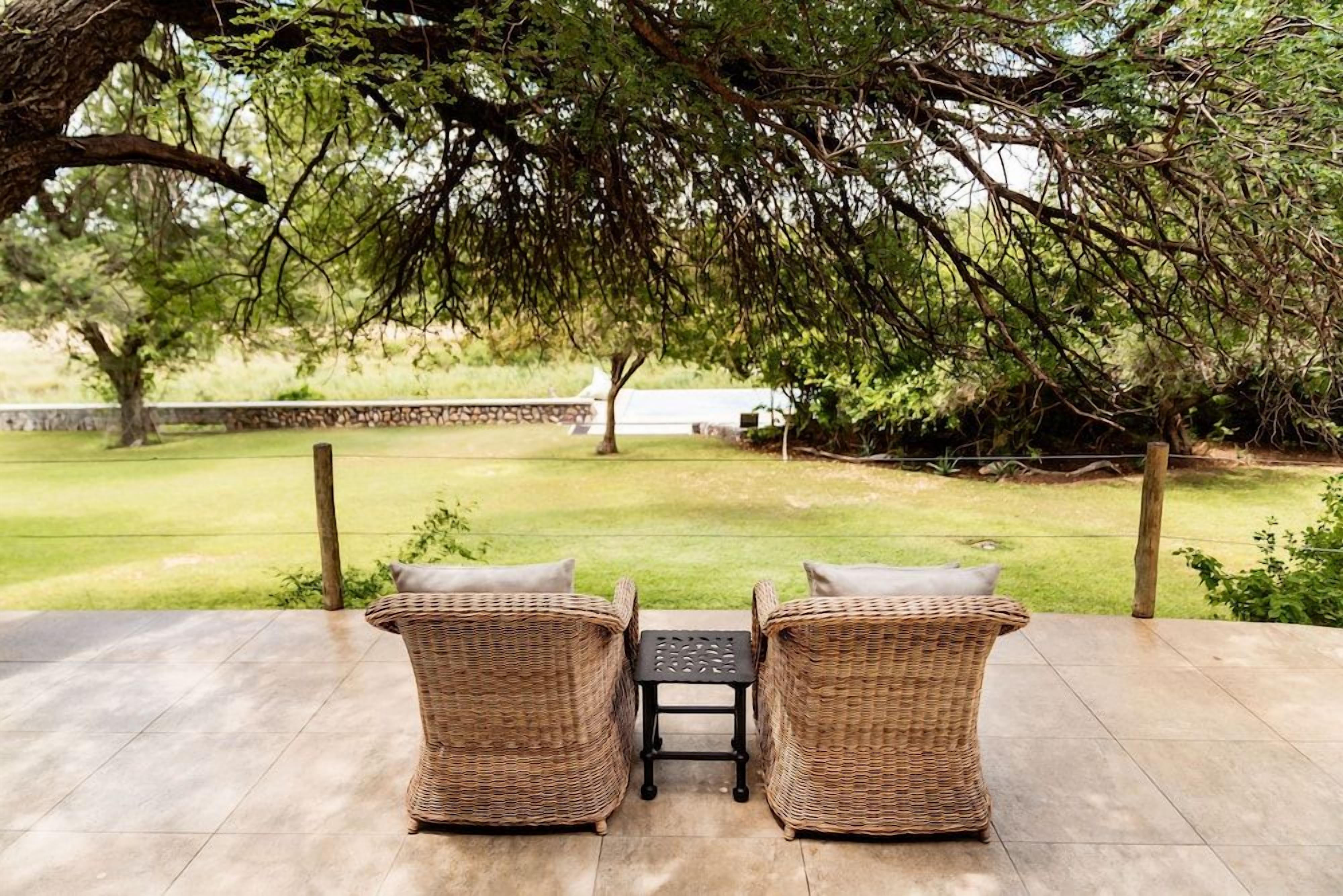 An outdoor patio with wicker furniture and a table, overlooking a grassy area with a tree and a stone wall in the background.