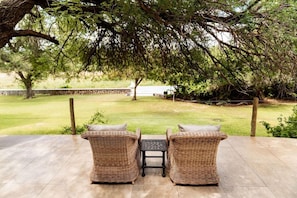 An outdoor patio with wicker furniture and a table, overlooking a grassy area with a tree and a stone wall in the background.