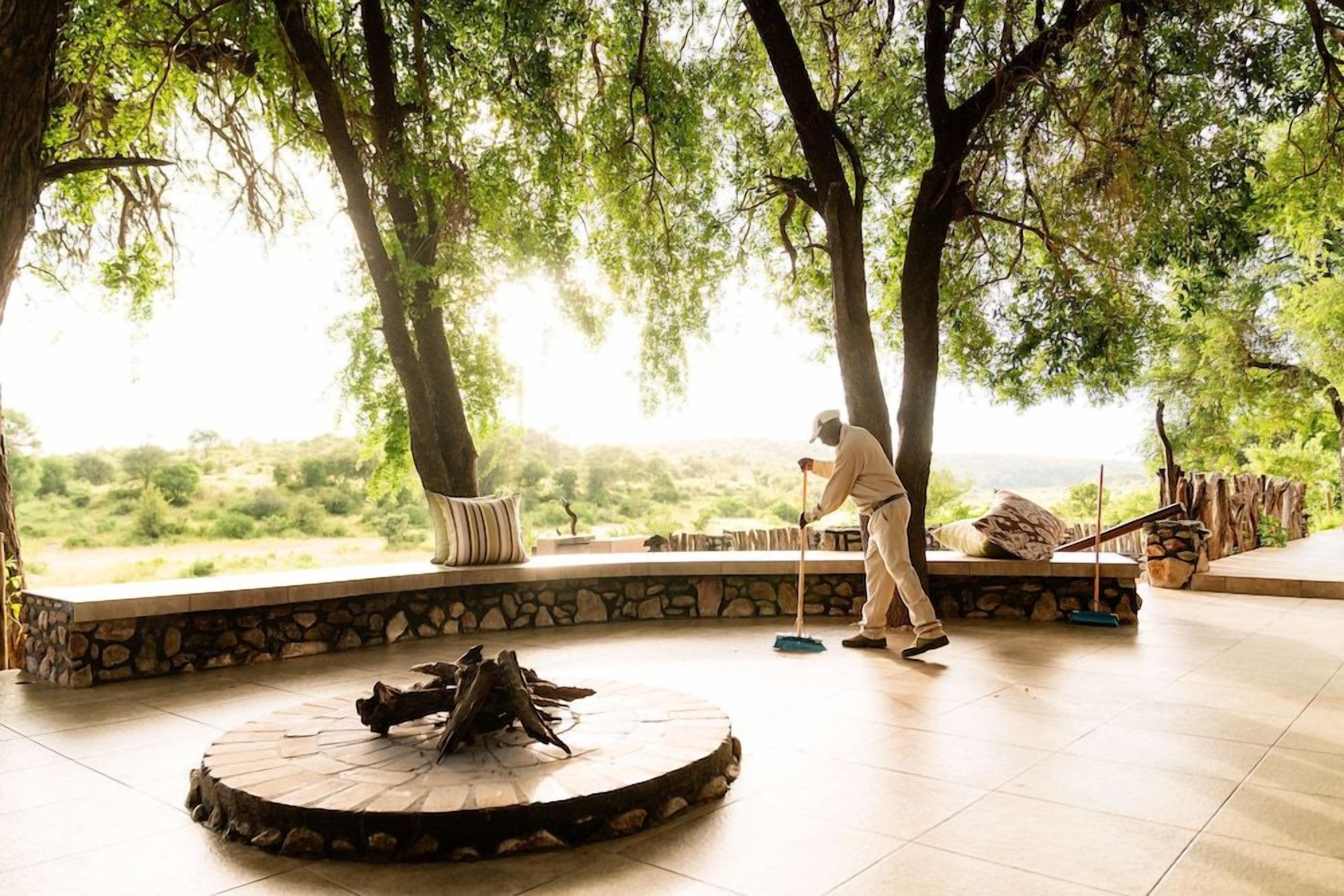 A person sweeping a stone-tiled patio with a wooden broom, surrounded by trees and a stone wall.