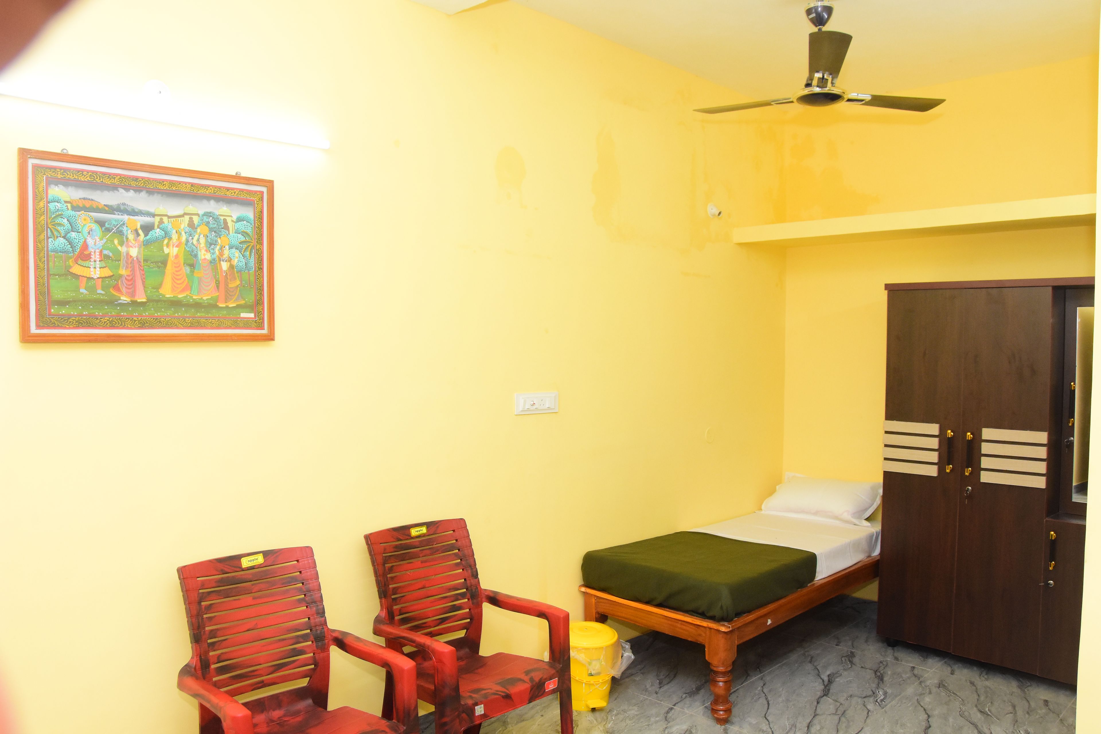 A room with a bed, two chairs, a ceiling fan, a wall-mounted light, and a framed picture.