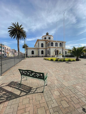 Una plaza con un banco, un edificio con torre del reloj y palmeras.