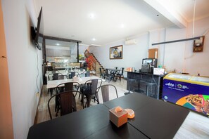A modern dining area with multiple tables and chairs, a kitchen in the background, and a staircase leading to an upper level. - Rehat at Zeva Guest House (Bandung)