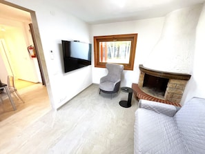 Ein Wohnzimmer mit Flachbildfernseher, einem grauen Sessel, einem kleinen Beistelltisch und einem Kamin.