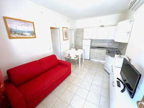A compact living space with a red sofa, a dining area, a kitchenette, and a TV. - Accommodation in Residence With Pool and Garden (San Michele al Tagliamento)