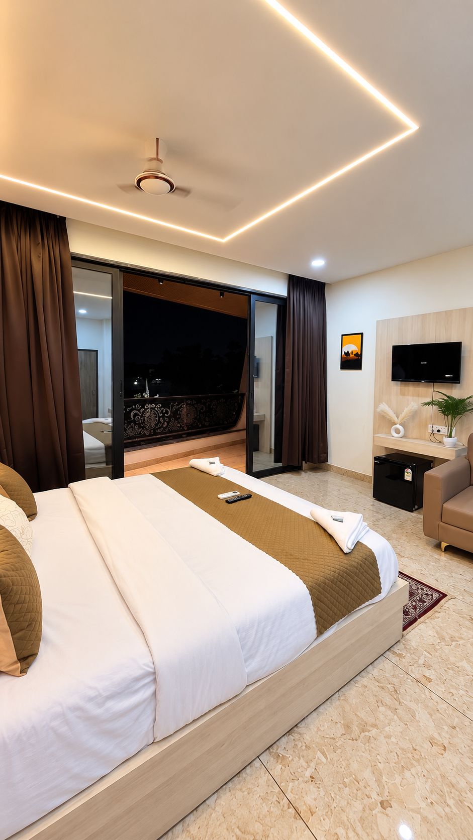 A modern hotel room with a large bed, a flat-screen TV, a desk, and a sliding glass door leading to a balcony.