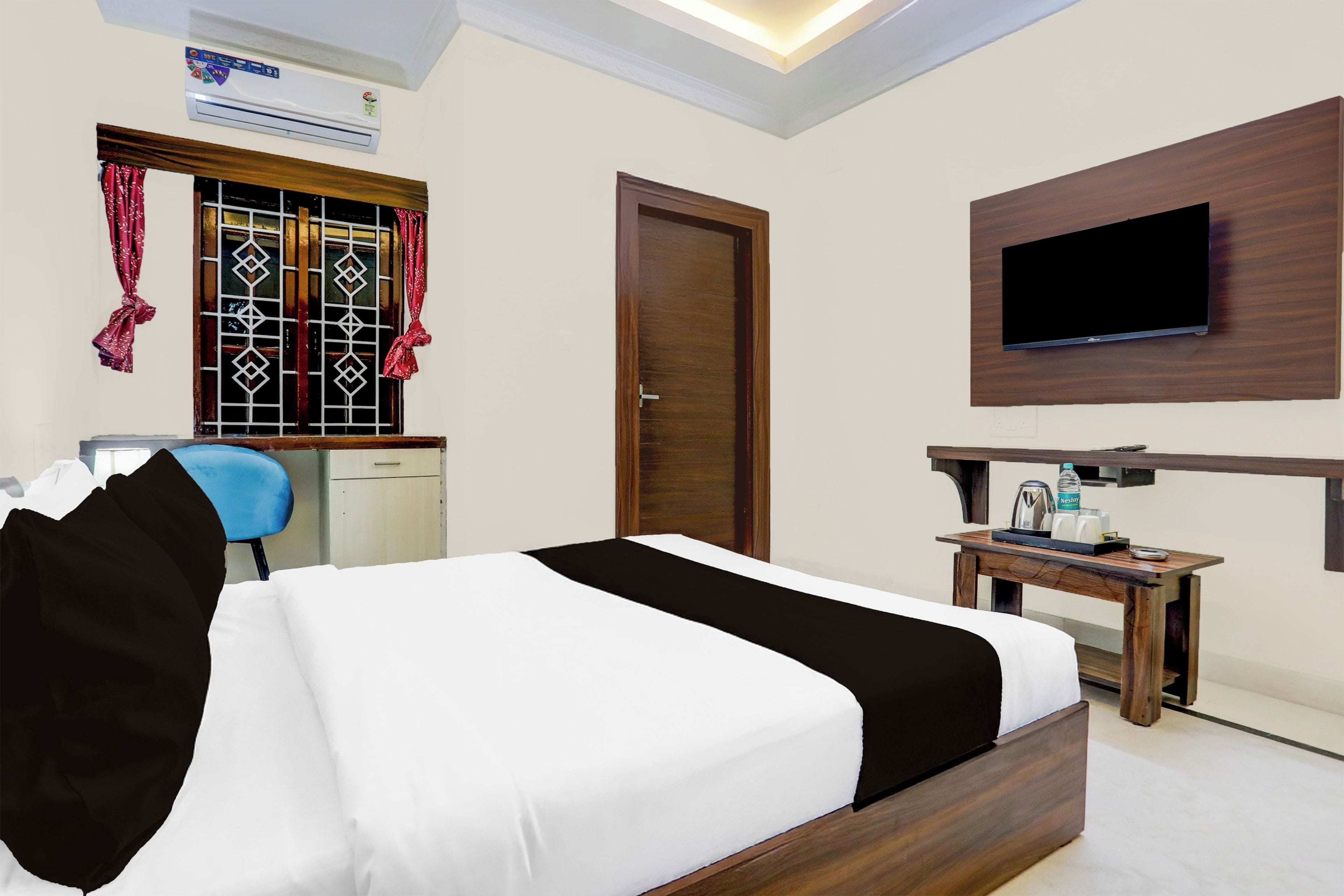 A hotel room with a bed, a desk, a chair, a TV mounted on the wall, and an air conditioning unit.