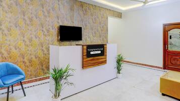 A reception area with a reception desk, a television mounted above it, a blue chair, and two potted plants.