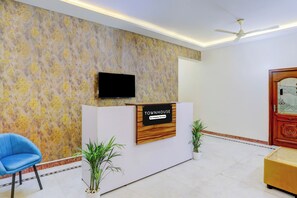 Un área de recepción con mostrador, televisor montado arriba, una silla azul y dos plantas en macetas.