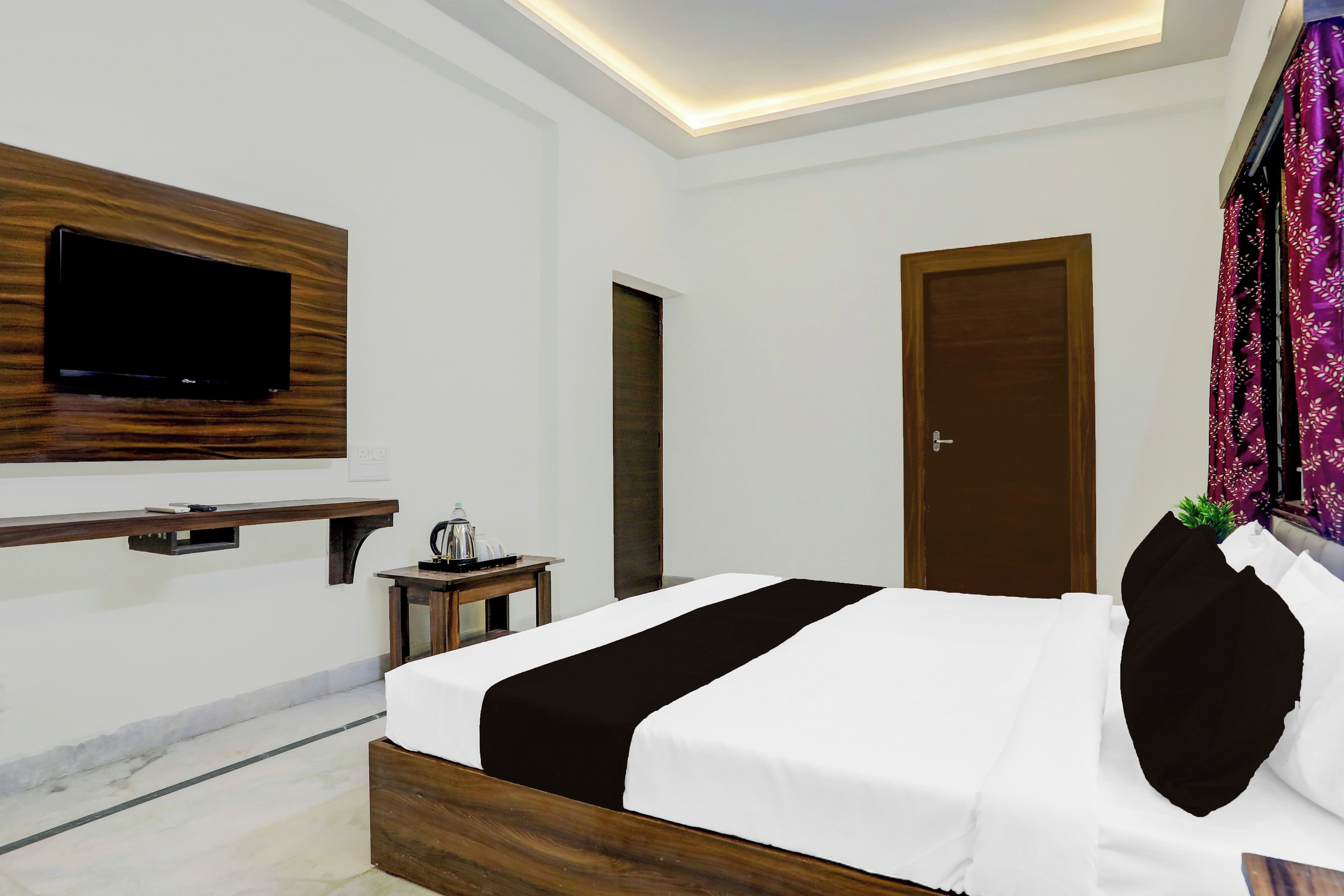 A modern hotel room with a bed, a TV mounted on the wall, a small table with a kettle, and a door.