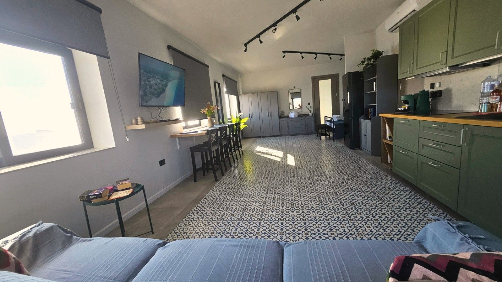 Ampla sala de estar com uma cama grande, mesa de jantar e cadeiras, cozinha com armários verdes e piso ladrilhado.
