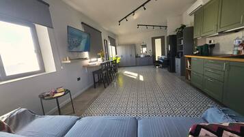 Ampla sala de estar com uma cama grande, mesa de jantar e cadeiras, cozinha com armários verdes e piso ladrilhado.