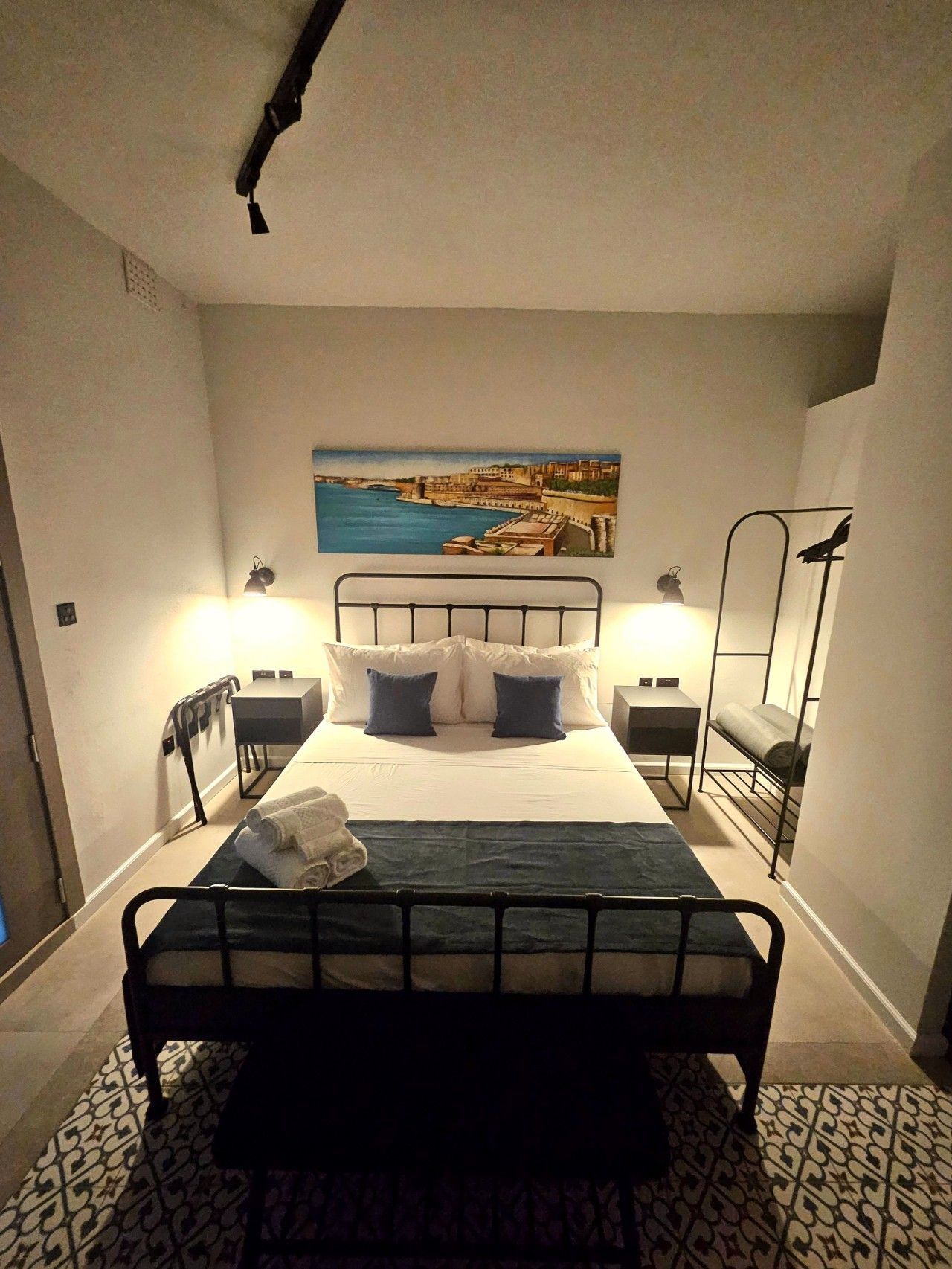 Une chambre à coucher avec un lit, des lampes de chevet, un tableau représentant un paysage côtier et un banc en métal.