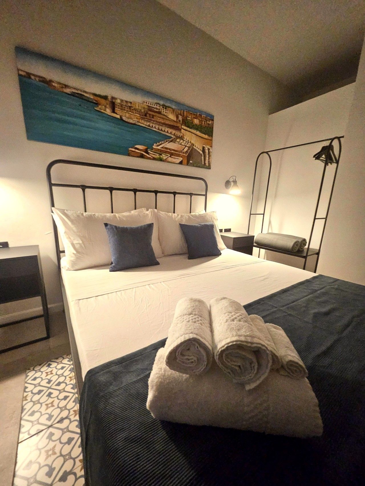 A hotel room with a bed, a painting of a coastal scene, a nightstand, a lamp, and a folded towel on the bed.