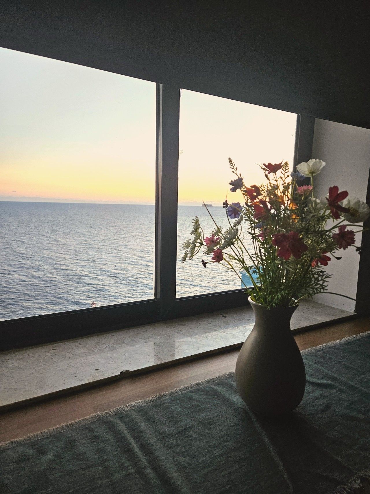 A room with a large window overlooking a body of water, a vase with flowers on a windowsill, and a textured rug on the floor.