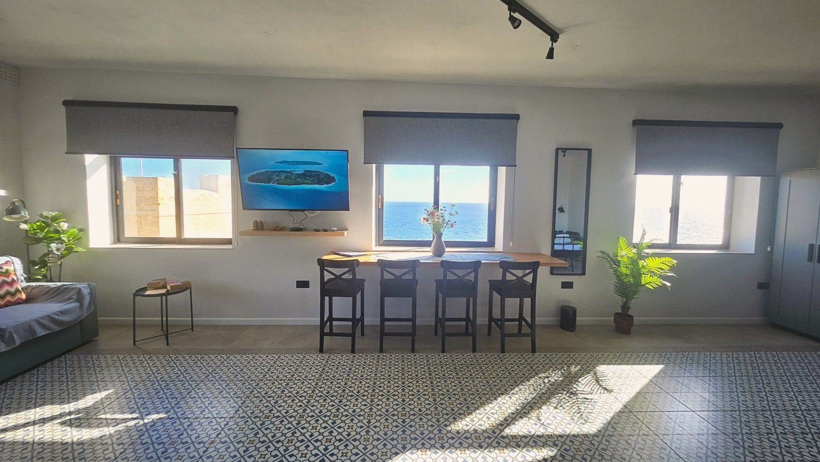 A spacious living area with a large window, a flat-screen TV, a dining table with chairs, and a patterned floor.