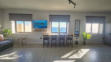 A spacious living area with a large window, a flat-screen TV, a dining table with chairs, and a patterned floor.