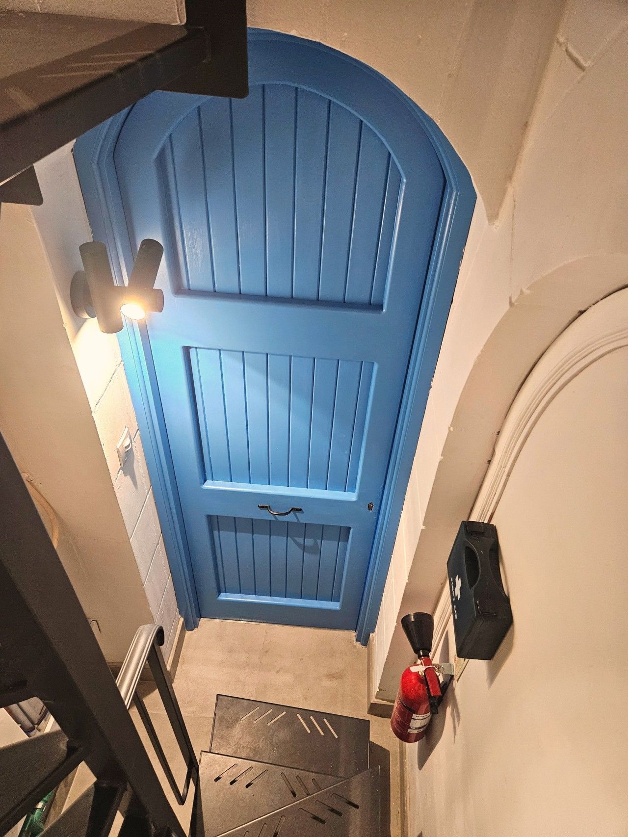 A blue door with a handle, a fire extinguisher mounted on the wall, and a metal grate on the floor.