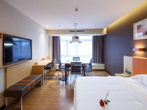 Habitación de hotel con cama, televisor, escritorio con sillas, ventana con cortinas y un banco.
