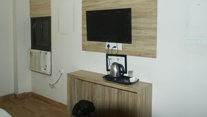 A room with a wooden cabinet, a mounted television, a window with blinds, and a wall-mounted air conditioning unit. - Hotel Shree Banaras (Varanasi)