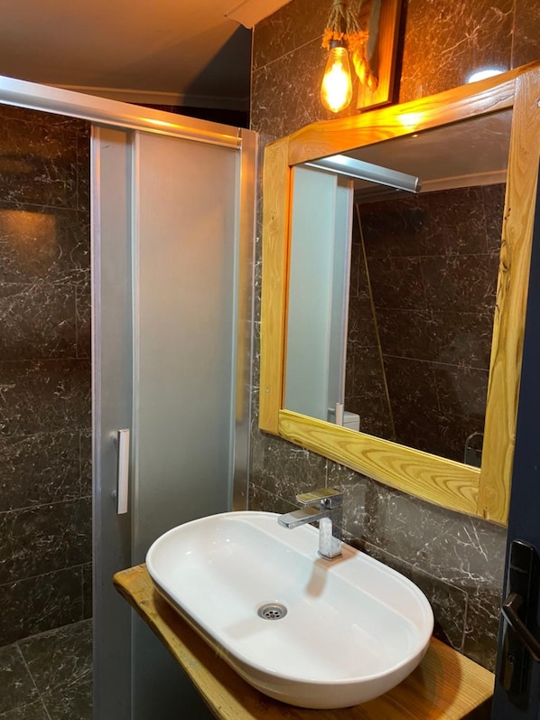 A bathroom with a white sink, a wooden vanity, a mirror with a wooden frame, and a shower area with a glass door. - Astroia Pansiyon (Lapseki)