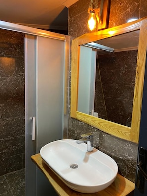 A bathroom with a white sink, a wooden vanity, a mirror with a wooden frame, and a shower area with a glass door. - Astroia Pansiyon (Lapseki)