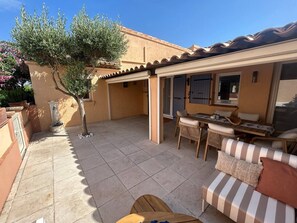 Een overdekt terras met een tafel, stoelen, een kleine boom en een bank met kussens.