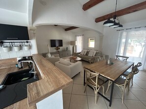 Een keuken met een houten aanrecht en een spoelbak, een eethoek met een houten tafel en stoelen, een bank en een televisie op de achtergrond.