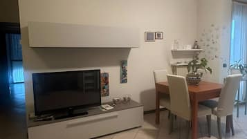Una sala moderna con un televisor de pantalla plana, una mesa de comedor con sillas y una zona de cocina con estantes.