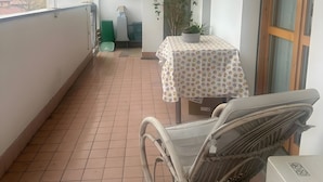 Een balkon met een betegelde vloer, een rieten stoel, een tafel met een tafelkleed met patroon, een potplant en een uitzicht op gebouwen in de verte.