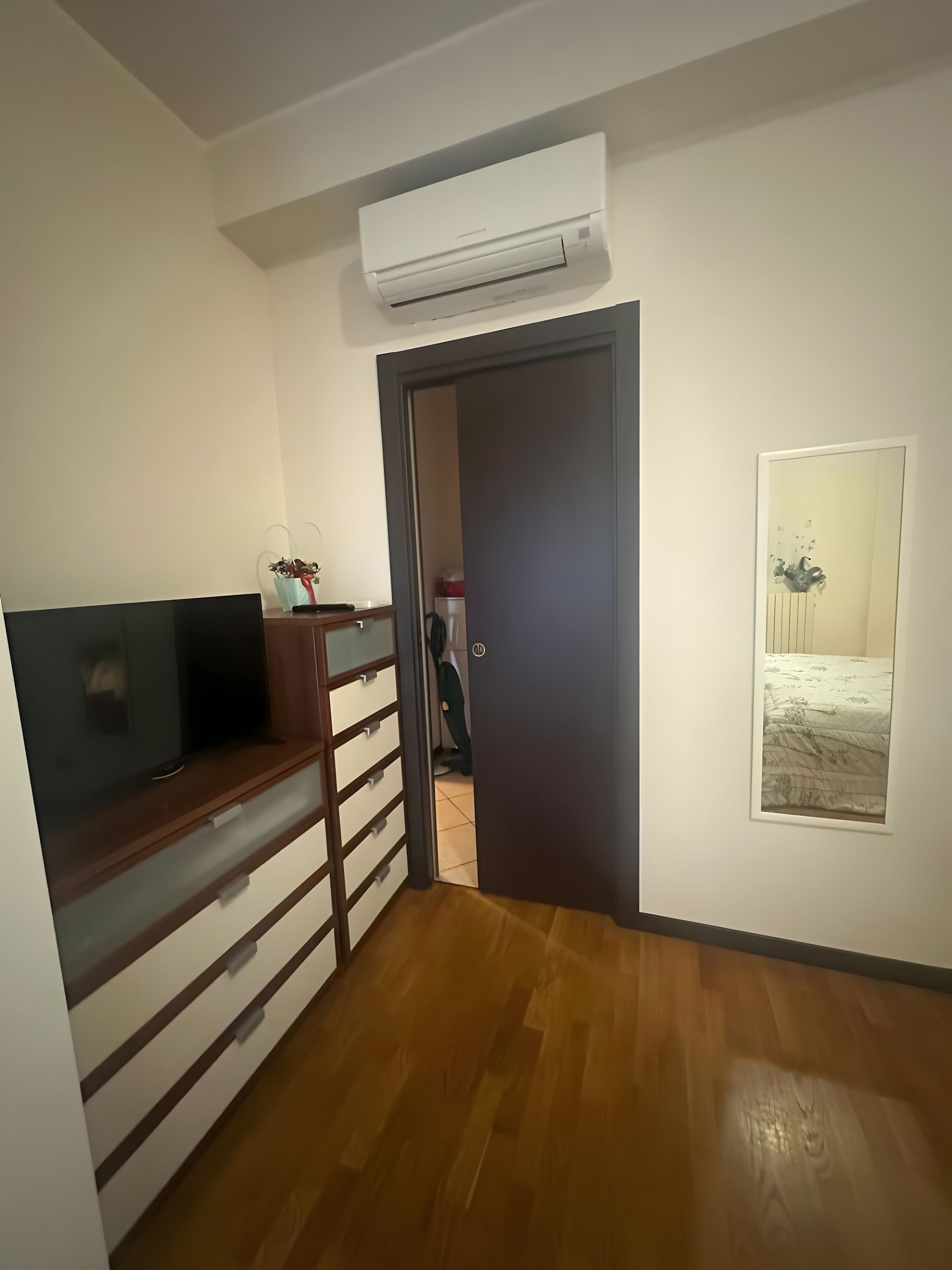Una habitación con piso de madera, un televisor sobre un mueble de madera, una puerta que da a otra habitación y un aire acondicionado instalado en la pared.