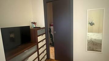 Una habitación con piso de madera, un televisor sobre un mueble de madera, una puerta que da a otra habitación y un aire acondicionado instalado en la pared.