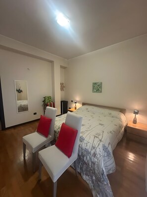 Un dormitori amb un llit gran, dues cadires blanques amb coixinets vermells, una petita taula de nit amb una làmpada i una obra d'art penjada a la paret.