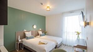 Habitación de hotel con dos camas, un escritorio y una silla. Hay un televisor montado en la pared, una mesita con un florero y una ventana con cortinas translúcidas.