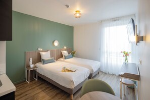 A hotel room with two beds, a desk, and a chair. There is a TV mounted on the wall, a small table with a vase of flowers, and a window with sheer curtains. - All Suites Appart Hotel Le Havre (Le Havre)