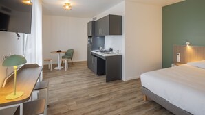 Una habitación de hotel compacta con kitchenette, una cama, un escritorio con lámpara y un pequeño comedor.