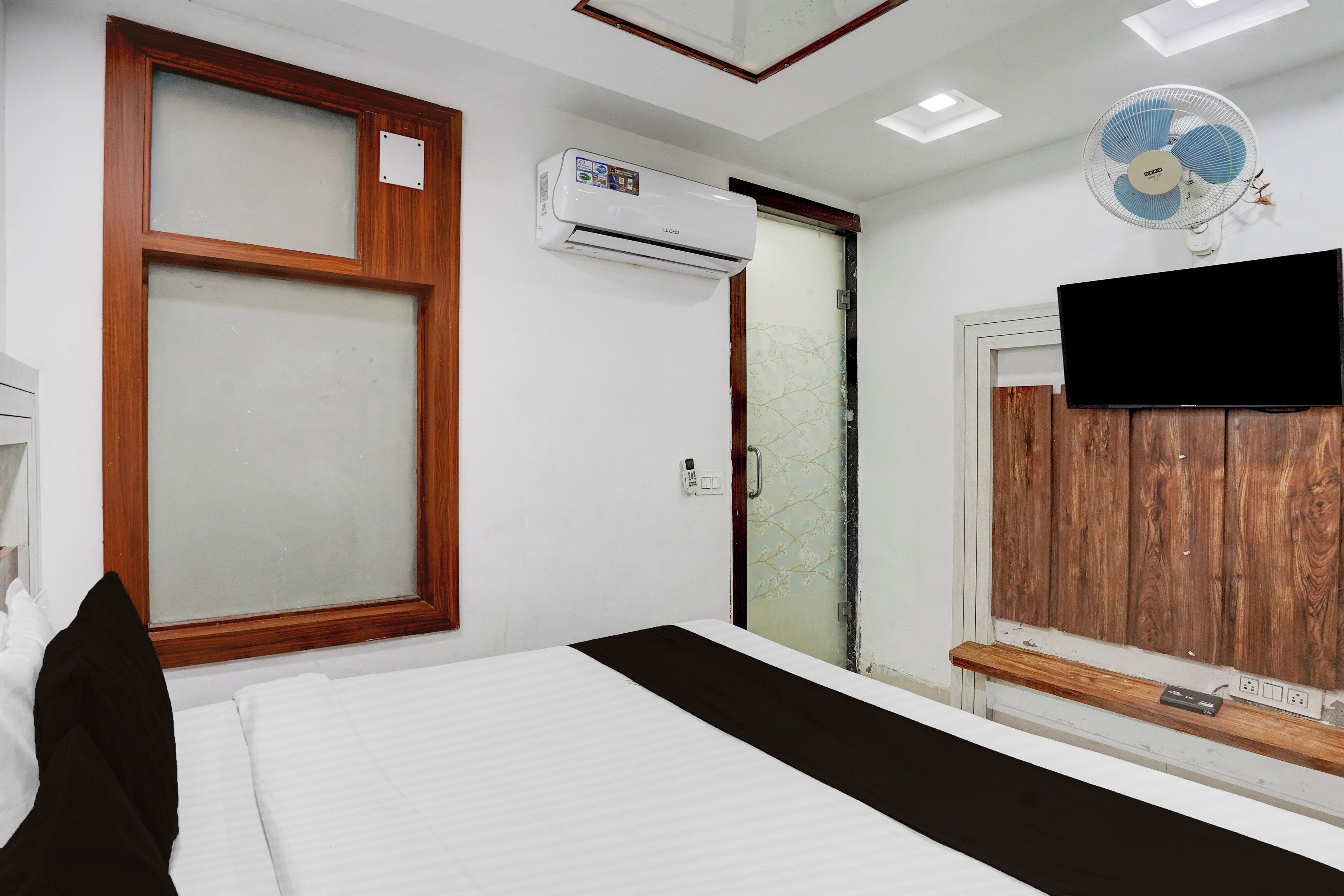 Habitación de hotel con cama, unidad de aire acondicionado, ventilador de pared, televisión y puerta con estampado floral.