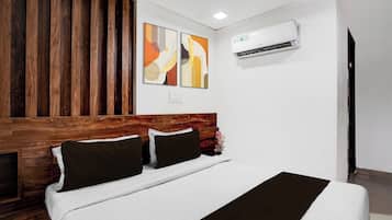 Habitación de hotel con cabecera de madera, cama con ropa de cama blanca y almohadas oscuras, dos cuadros abstractos en la pared y un equipo de aire acondicionado instalado en la pared.