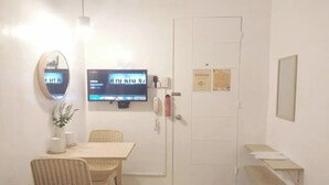 A room with a television mounted on the wall, a door with a sign, a small table with a chair, and a trash bin. - VG Pension and Residences by Zuzu (Cebu City)